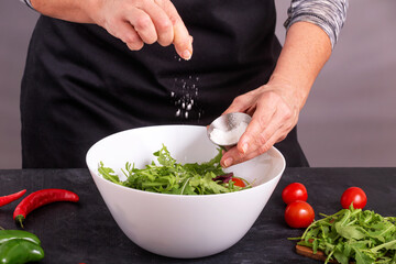 A cook sprinkles salt on a fresh vegetable salad with tomatoes and arugula, closeup with selective focus