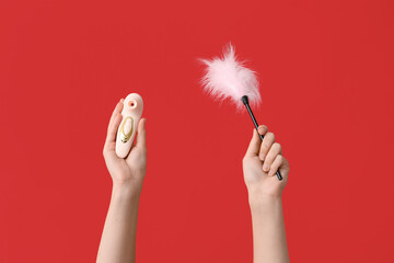 Female hands with vibrator and feather stick on red background, closeup - obrazy, fototapety, plakaty