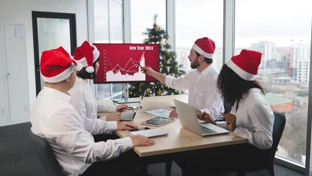 Successful team of multiethnic business people in Santa hats raising hands and screaming with happiness while working together in decorated with Christmas tree office in business center.