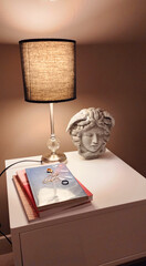 Mesa de noche con lámpara cálida, libros y una estatua de la cabeza de la gorgona en piedra