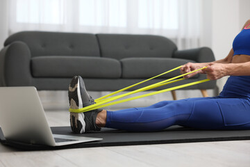 Woman doing exercise with fitness elastic band near laptop on mat at home, closeup