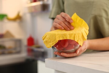 Woman covering bowl of fresh tomatoes with beeswax food wrap at table in kitchen, closeup. Space...