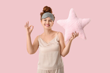 Beautiful young woman in sleep mask holding soft pillow in shape of star and showing ok gesture on pink background