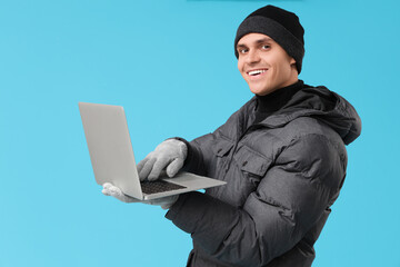 Young man in stylish puffer jacket with modern laptop on blue background