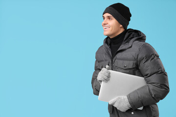 Young man in stylish puffer jacket with modern laptop on blue background