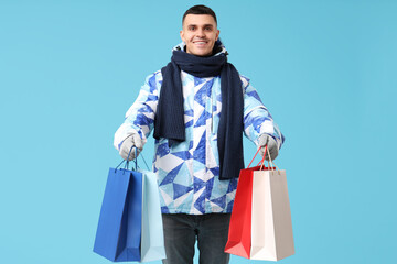 Young man in stylish puffer jacket and scarf with shopping bags on blue background
