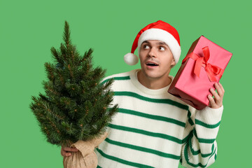 Young man in Santa hat with Christmas tree and gift box on green background