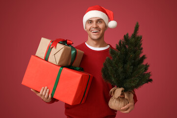 Young man in Santa hat with Christmas tree and gift boxes on red background