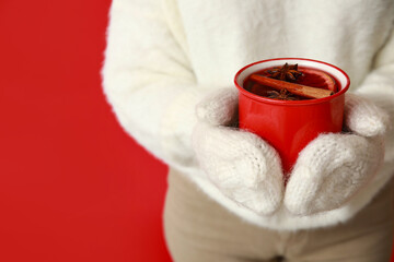 Young woman in warm mittens with cup of hot mulled wine on red background