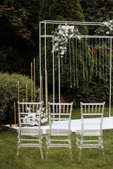 A beautiful wedding arch decorated with flowers, electric hanging lamps and a row of chairs stands on the green grass of a lawn in a garden, park. 