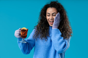 Beautiful young African-American woman in gloves with glass cup of hot mulled wine on blue...