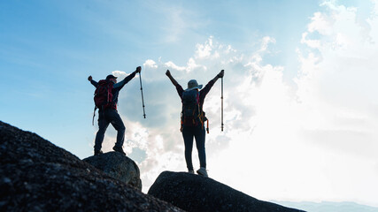 Silhouette of Asian Male and female standing raised hands with trekking poles on cliff edge on top...
