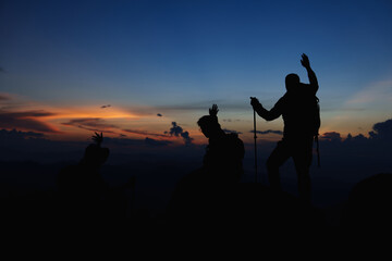 Fototapeta na wymiar Silhouette of Asian teamwork standing raised hands with trekking poles and Backpack and other camping gear on cliff edge on top of rock mountain with at sunset rays over the clouds background, 
