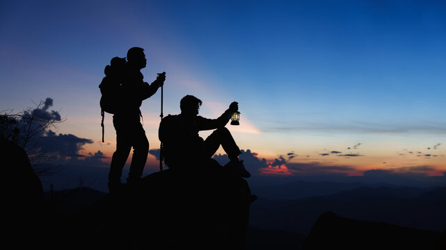 Silhouette of Asian two male standing raised hands with trekking poles and kerosene black lamp on cliff edge on top of rock mountain with at sunset rays over the clouds background, 