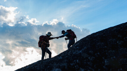 Silhouette of Asian Male and female hikers climbing up mountain cliff and one of them giving helping hand with friend at sunset, People helping, Asia couple hiking help each other concept.