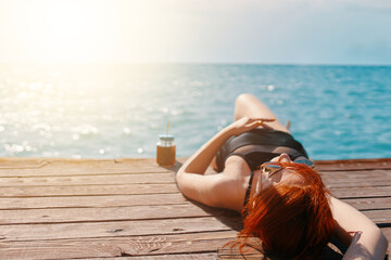 Red-haired girl is enjoying a vacation at the sea. Woman in a swimsuit is lying on a wooden pier with a bottle of lemonade and sunbathing. Summer trip. Copy space for travel agency advertising.