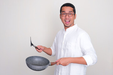 Adult Asian man showing wow expression while holding empty frying pan and spatula