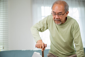 Elderly man suffering from knee pain ache holding handle of cane, senior old man typing to stand up from sofa alone with walking cane stick to walk at home, retirement medical healthcare concept
