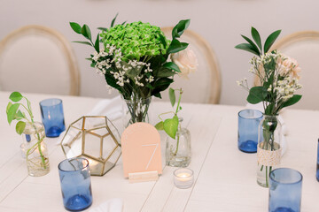 A modern table surrounded by chairs, and decorated with greenery in vases, glass geometric shapes and blue candleholders on white tablecloth