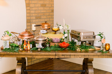 A beautiful decorated serving table with food in metal and gold containers. The table is decorated with flowers and greenery and candles. A beautiful wedding reception brunch 