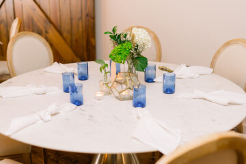 An elegant decorated dining table with a simple centerpiece with flowers and greenery and blue candleholders, and white napkins on the table