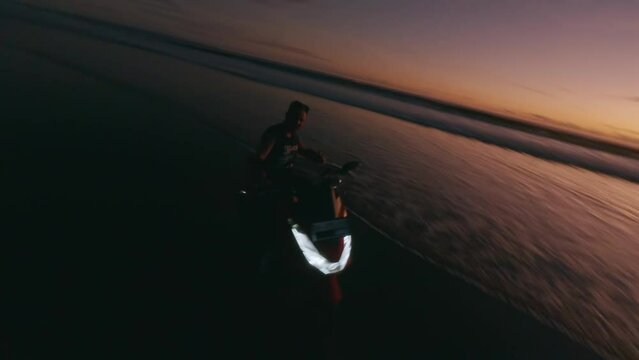 Aerial view man motorcyclist riding red bike one hand shaka night sunset beach sea tropical landscape. FPV sport drone cinematic shot evening dusk coast flying over endless tide calm foam wave ocean