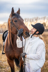 Fototapeta premium Beautiful blond professional female jockey standing near horse in field. Friendship with horse concept