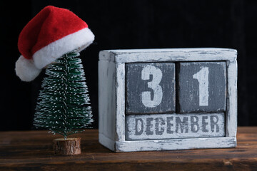 December 31 on wooden calendar standing next to Christmas tree decorated with santa claus hat. - obrazy, fototapety, plakaty