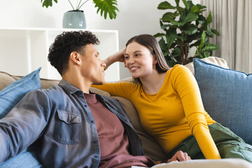 Fototapeta premium Happy diverse couple sitting on couch and looking at each other in living room at home, copy space