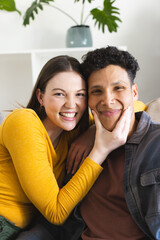 Happy diverse couple sitting on couch and holding face in hand in living room at home, copy space
