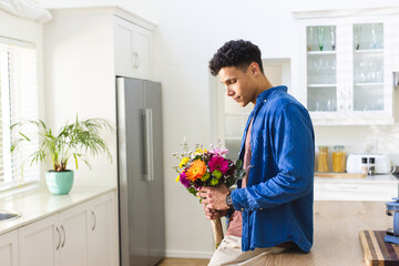 Happy biracial man holding flowers in kitchen at home, copy space - Powered by Adobe