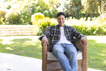 Portrait of happy biracial man sitting in chair in sunny garden, copy space