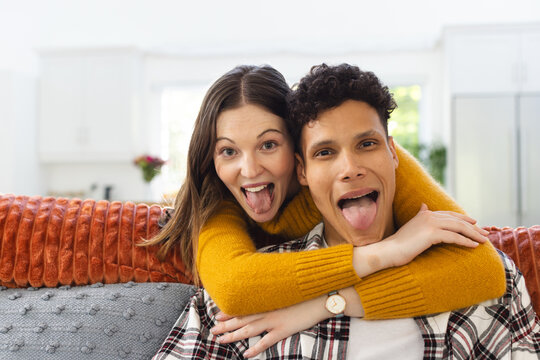 Portrait of happy diverse couple embracing on couch and making funny faces at home, copy space