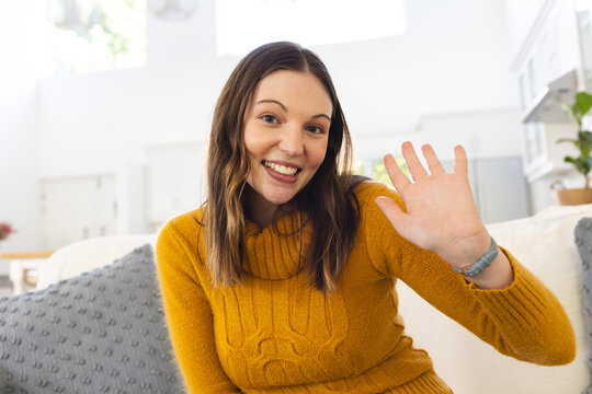 Portrait of happy caucasian woman sitting on couch and waving with hand at home, copy space