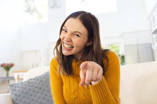 Portrait of happy caucasian woman sitting on couch and pointing with finger at home, copy space