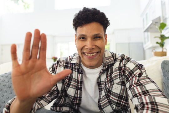 Portrait of happy biracial man sitting on couch and waving with hand at home, copy space