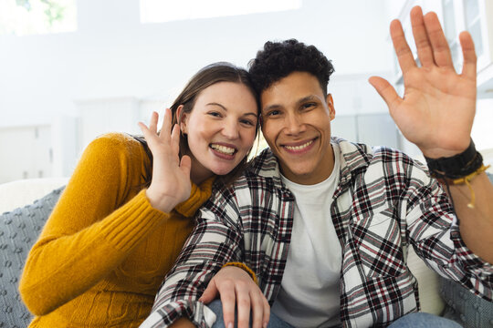 Portrait of happy diverse couple sitting on couch, embracing and waving hands at home, copy space
