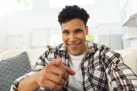 Portrait of happy biracial man sitting on couch and pointing with finger at home, copy space