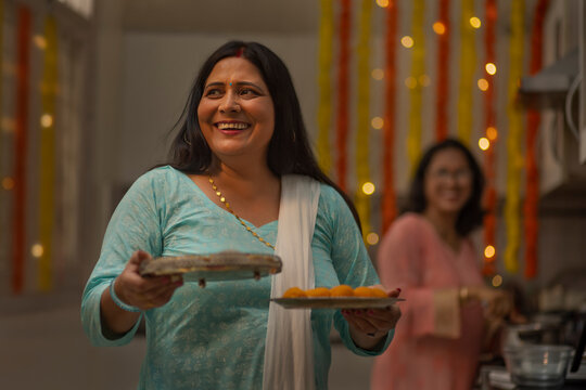 Middle-aged woman holding plates of sweets while standing in the kitchen on the occasion of Diwali