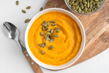 Above close up view of homemade vegetarian pumpkin soup made of pureed vegetables with creamy texture decorated with dried seeds or pepita served in bowl with spoon and on wooden board on white table