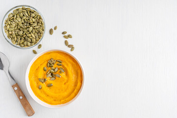 Top view of creamy homemade traditional pumpkin soup made of pureed squash decorated with seeds or pepita served with spoon on white wooden background with copy space for healthy vegetarian dinner
