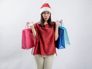 A portrait of a happy Asian woman celebrating Christmas, wearing a red dress and Santa hat, carrying a shopping bag. Isolated against a white background.