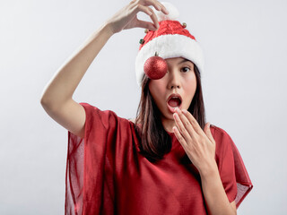 A portrait of a joyful Asian woman celebrating Christmas, dressed in a red outfit and Santa hat, holding Christmas accessories like ornaments and playing with them. Isolated against a white background