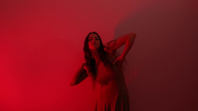 Beautiful sexy woman wearing dress dancing in red lighted room. Sensual scene of female model performing in front of the wall in night