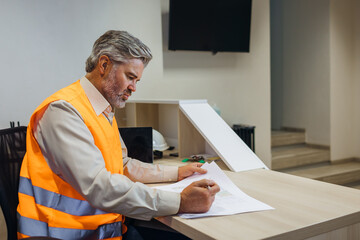 builder, engineer in reflective vest uniform on desk in office looking through papers or sketches.