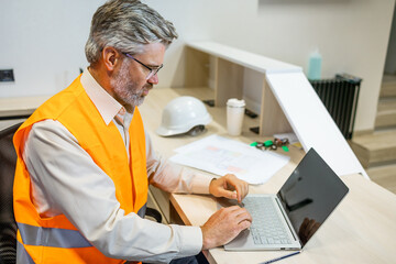 mature senior male sitting and using laptop at office factory. man worker in vest safety typing - obrazy, fototapety, plakaty