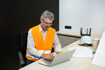 senior mature man engineer with laptop computer typing on laptop wear safety vest. Work housing...
