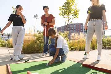 Cute school girl playing mini golf with family. Happy toddler child having fun with outdoor activity. Summer sport for children and adults, outdoors. Family vacations or resort.
