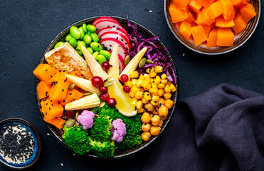 Vegan buddha bowl with sweet potato, quinoa, chickpeas, soybeans edamame, tofu, corn, cabbage, radish, broccoli and seeds, black table background, top view. Autumn or winter healthy vegetarian food