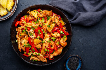 Stir fry chicken with pineapple, red paprika, chives, soy sauce and sesame seeds in frying pan. Asian cuisine dish. Black table background, top view